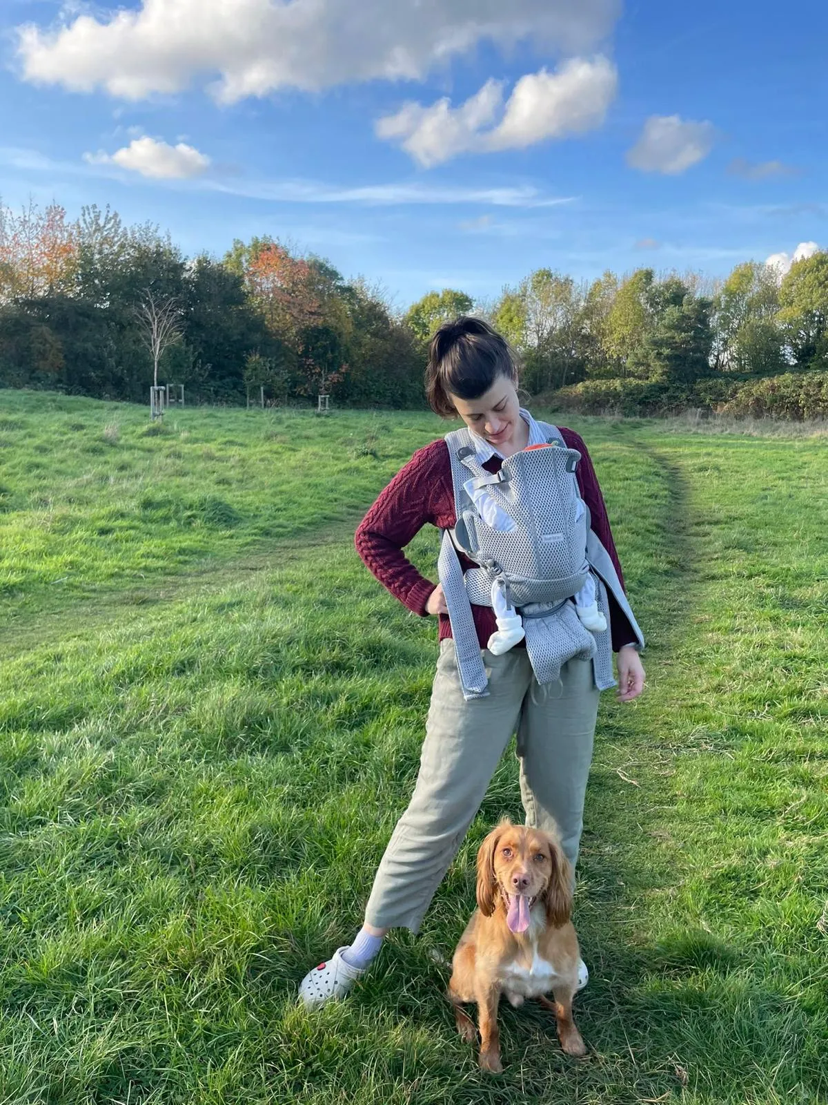 Georgia standing in a field on a bright autumn day, her baby in a carrier and her spaniel at her feet